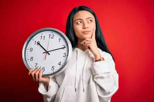 Young beautiful chinese woman holding big clock standing over isolated red background serious face thinking about question, very confused idea